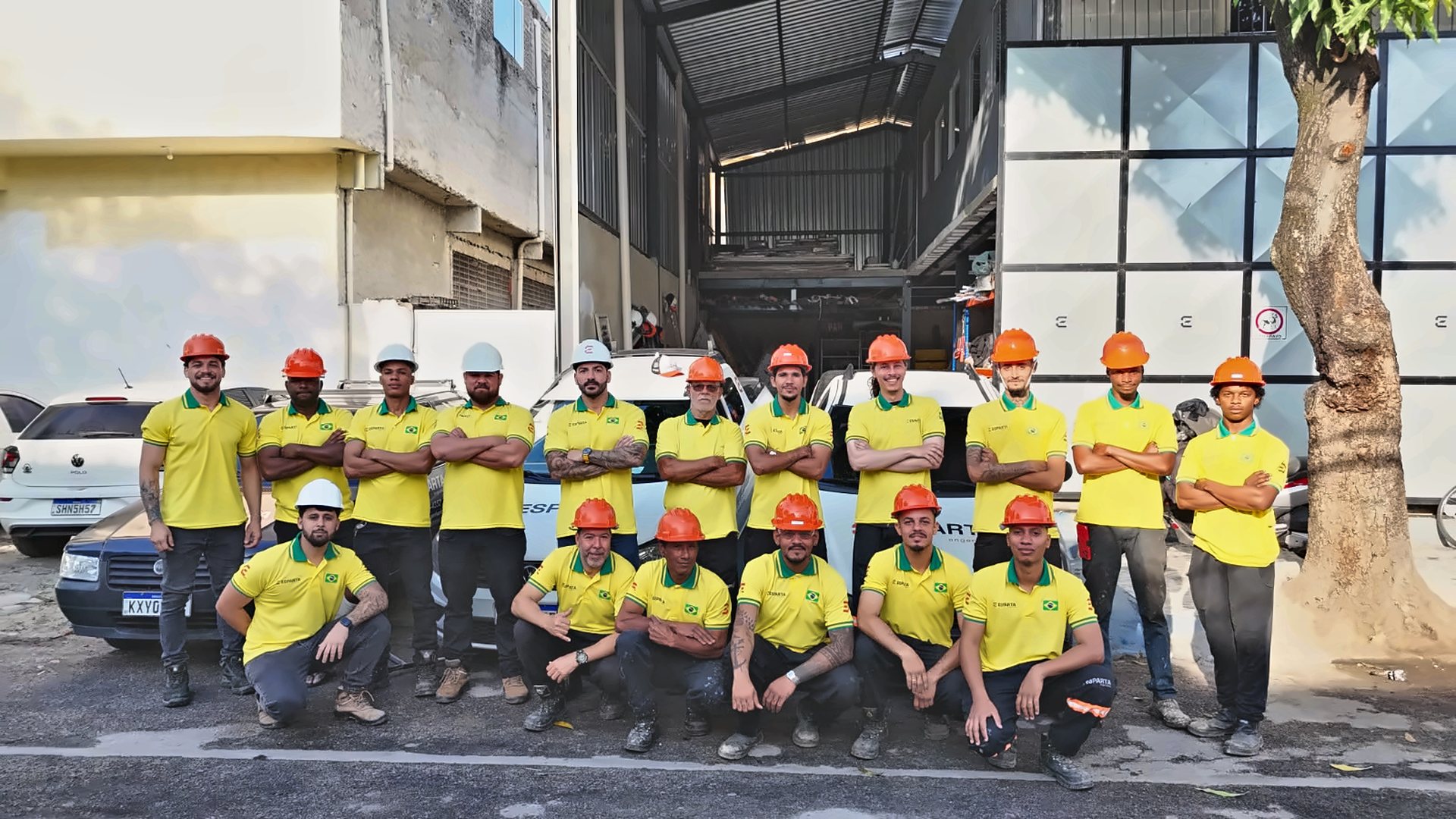 Equipe da Esparta Engenharia reunida de camisa amarela e capacete na frente da sede, com os carros da empresa ao fundo