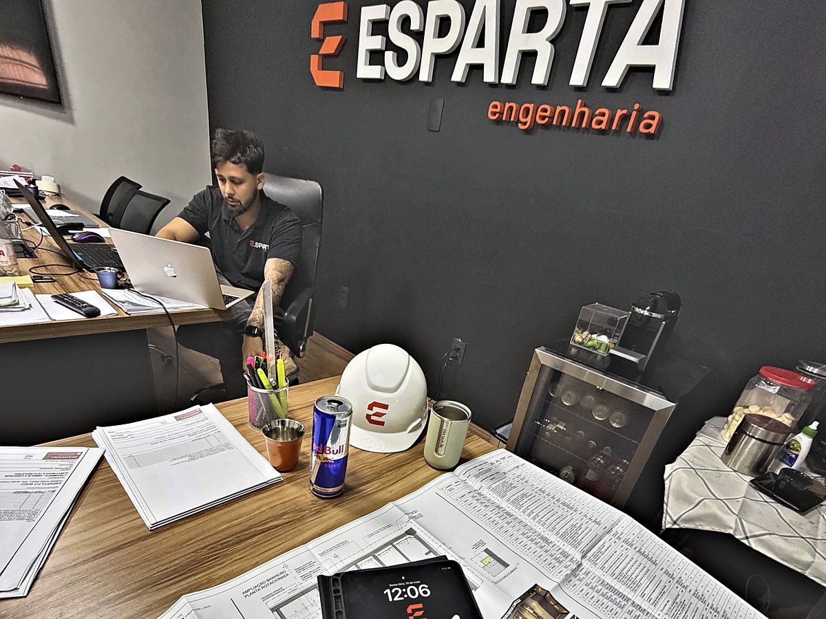 Engenheiro da Esparta Engenharia no escritório, com as plantas do projeto abertas na mesa e o logo da empresa na parede
